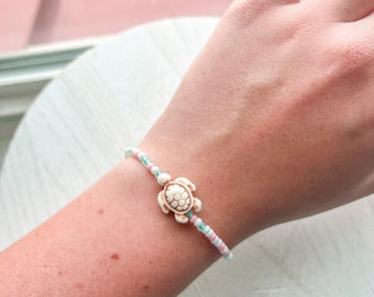 Puede incluir: Un brazalete de cuentas blancas y azul claro con un pequeño dije de tortuga blanca. El brazalete está en la muñeca de una persona.