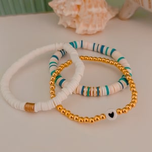 May include: Three bracelets stacked on a white surface. The top bracelet is white with blue and gold accents. The middle bracelet is white with blue and gold accents. The bottom bracelet is gold with a white bead with a black heart.
