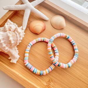 Puede incluir: Dos pulseras coloridas hechas de cuentas redondas de arcilla apiladas. Las pulseras son de color rosa, azul, naranja y blanco. Las pulseras están en una bandeja de madera con conchas marinas y una estrella de mar.