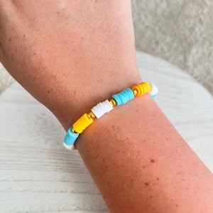 Puede incluir: Un brazalete de cuentas coloridas con cuentas amarillas, blancas y azules. El brazalete está hecho de cuentas pequeñas y redondas que están enhebradas en una cadena dorada.