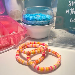 Puede incluir: Tres pulseras coloridas hechas de cuentas redondas de plástico apiladas. Las pulseras son de color rosa, naranja y amarillo.