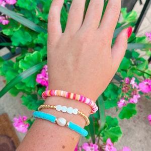 Puede incluir: Una mano con tres pulseras. La pulsera superior es rosa, naranja y blanca. La pulsera del medio es dorada con cuentas blancas. La pulsera inferior es turquesa con un dije de perla blanca.