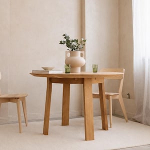 Könnte beinhalten: Ein runder Esstisch aus hellem Holz mit vier geraden Beinen, begleitet von zwei passenden Holzstühlen. Auf dem Tisch steht eine große beige Keramikvase mit zwei Henkeln, die grünes Blattwerk enthält, eine kleine Schale und zwei grüne Gläser.