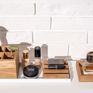 Könnte beinhalten: Holzorganizer mit Fächern für Make-up-Pinsel, einen Jade-Roller und Flaschen mit Foundation und Concealer. Der Organizer steht auf einer weißen Arbeitsplatte vor einer weißen Backsteinwand. Der Text "MAKE UP FOR EVER" ist auf einer Kompaktdose sichtbar.