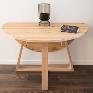 Op de afbeelding: Een lichtgekleurde houten halve cirkel tafel met een centrale steun en schuine poten. Een zwarte metalen kandelaar en een boek liggen op de tafel. De tafel staat op een donkere houten vloer.