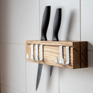 Op de afbeelding: Een houten messenhouder met vier witte haken en twee zwarte messen. De houder is gemonteerd op een witte betegelde muur.