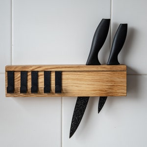 Op de afbeelding: Een houten messenhouder met zwarte messen. De houder is gemaakt van lichtgekleurd hout en heeft een reeks zwarte haken. Twee messen zijn in de houder gestoken en de lemmeten zijn zichtbaar. De messen hebben zwarte handvatten.