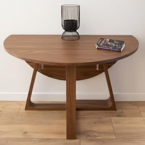 May include: A walnut-finished, semi-circular table with a central support and curved base. A black metal candle holder and a book are placed on the table. The table is set against a white wall and a wooden floor.
