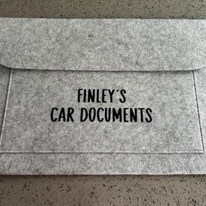 May include: A grey felt document holder with a flap closure. The text "FINLEY'S CAR DOCUMENTS" is printed in black on the front. Designed for storing car documents, it's a practical accessory.