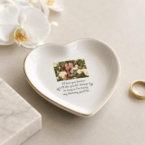 May include: A white heart-shaped ceramic trinket dish with a gold-toned rim. The center features a printed photograph of two people among yellow flowers, with the text: I'll love you forever. I'll like you for always. As long as I'm living my Mommy you'll be.