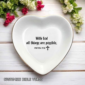 May include: White heart-shaped trinket dish with a gold rim. The dish features the text "With God all things are possible" and a cross symbol. The words "Customize Bible Verse" are at the bottom.