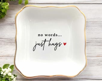 May include: A white ceramic trinket dish with a gold rim and a wavy edge. The dish features the words "no words... just hugs" in black script, with a small red heart. The dish is on a light wood surface.