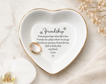 Könnte beinhalten: Eine weiße, herzförmige Keramikschale mit goldenem Rand. In der Mitte steht in schwarzer Schrift: Friendship, Friends give hope when life is low, Friends are a place where we can go, Friends are precious, honest & true, I feel so lucky that my friend is you, ergänzt durch zwei kleine Gänseblümchen und ein Herzsymbol. Ein schlichter goldfarbener Ring liegt in der Schale.
