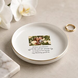 May include: A round white ceramic trinket dish with a gold-colored rim. The center features a printed photograph of a person and a child among yellow flowers, with the text: I'll love you forever. I'll like you for always. As long as I'm living my Mommy you'll be.