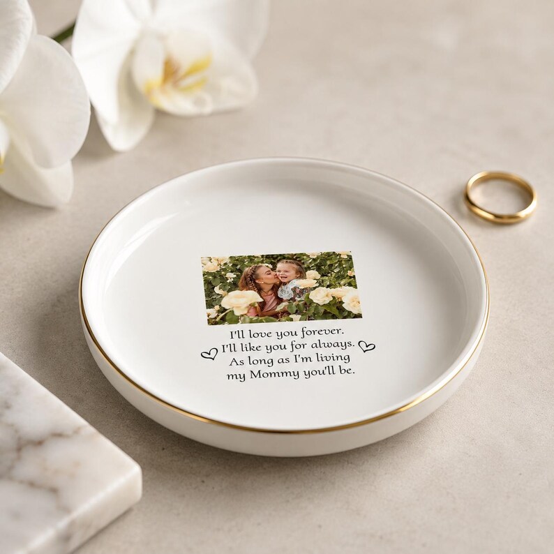 May include: A round white ceramic trinket dish with a gold-colored rim. The center features a printed photograph of a person and a child among yellow flowers, with the text: I'll love you forever. I'll like you for always. As long as I'm living my Mommy you'll be.