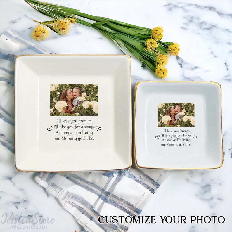 May include: Two square white ceramic trinket dishes with gold-toned rims. Each dish features a printed photograph of a person kissing a child in a garden, accompanied by the text: I'll love you forever. I'll like you for always. As long as I'm living my Mommy you'll be.