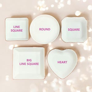 Puede incluir: Cinco bandejas de cer&aacute;mica blanca para joyas con ribete dorado. Las bandejas tienen forma de cuadrado, c&iacute;rculo, coraz&oacute;n y dos cuadrados de diferentes tama&ntilde;os. Las bandejas est&aacute;n etiquetadas como "LINE SQUARE", "ROUND", "SQUARE", "BIG LINE SQUARE" y "HEART".