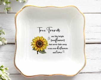 May include: A square white ceramic trinket dish with scalloped edges and a gold-painted rim. The centre features a yellow sunflower illustration and black text that reads: True Friends are like bright sunflowers that never fade away even over distance and time.