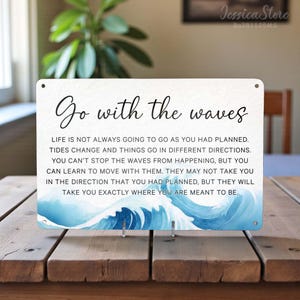 May include: A rectangular metal sign with the text "Go with the waves" in black script. The sign includes a watercolor image of a blue wave and inspirational text. The sign is displayed on a wooden table.