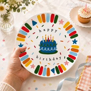 Puede incluir: Un plato de cerámica blanca con una ilustración central de un pastel de cumpleaños azul con cinco velas encendidas. El texto "happy birthday" está impreso en negro. El borde está decorado con pinceladas coloridas, estrellas y triángulos.