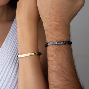 May include: Two bracelets with black beaded bands and metal bars. One bar is gold-toned with the engraved date 01.03.2020, and the other is black with the engraved Roman numeral date XXVI.XII.MMXX. Both bracelets feature a sleek, minimalist design.