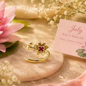 Può includere: Un anello aperto color oro con un design floreale e una piccola pietra rossa al centro. Accanto all'anello si trova un cartoncino rettangolare con il testo July, Ruby & Water Lily, Passion, Positivity, Strength e un'illustrazione di ninfea.