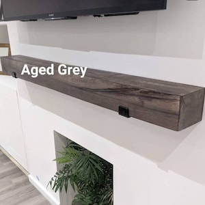 May include: Aged grey floating mantel shelf. The rectangular shelf is mounted on a white wall with black metal brackets. The text "Aged Grey" is displayed on the shelf.