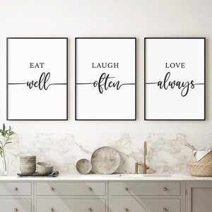 May include: Three black framed prints with white backgrounds and black calligraphy. The prints read "Eat well", "Laugh often", and "Love always". The prints are hanging on a white wall above a grey kitchen cabinet.