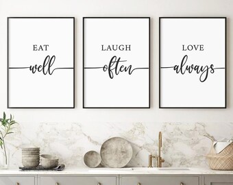 May include: Three black framed prints with white backgrounds and black calligraphy. The prints read "Eat well", "Laugh often", and "Love always". The prints are hanging on a white wall above a gray kitchen cabinet.