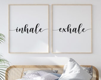 May include: Two framed prints with the words "inhale" and "exhale" in black cursive script on a white background. The prints are hanging on a white wall above a bed with white bedding.