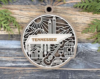 May include: Wooden ornament featuring a detailed laser-cut design of Tennessee landmarks. The design includes mountains, a sun, the state name, a guitar, and a barrel. The ornament is round with a hole for hanging and is made of layered wood.