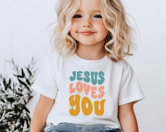 May include: A white t-shirt featuring the text "JESUS LOVES YOU" in teal, coral, and gold. The shirt is worn by a young child with blonde hair.