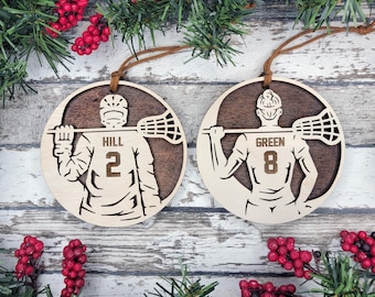 May include: Two wooden lacrosse ornaments. Each ornament is circular with a light wood cutout of a lacrosse player holding a lacrosse stick. One ornament has the name "Hill" and the number "2", the other has "Green" and "8". Both have brown leather hanging strings.
