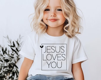 May include: A white t-shirt featuring the text "JESUS LOVES YOU" within a black square frame, with a small black heart. The t-shirt is worn by a young child with blonde hair.
