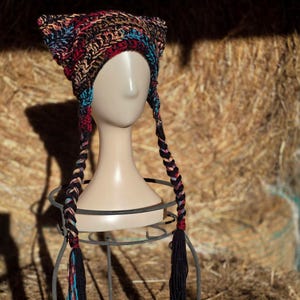 May include: A handmade crochet hat with cat ears, displayed on a mannequin head. The hat features a mix of colors including black, red, blue, and tan. Long, braided tassels hang down on either side, adding a playful touch to this unique accessory.