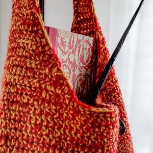 May include: A handmade crochet bag in shades of red and gold, with a long strap. The bag contains a book with a pink cover and a black wand. The bag is made of a textured yarn.