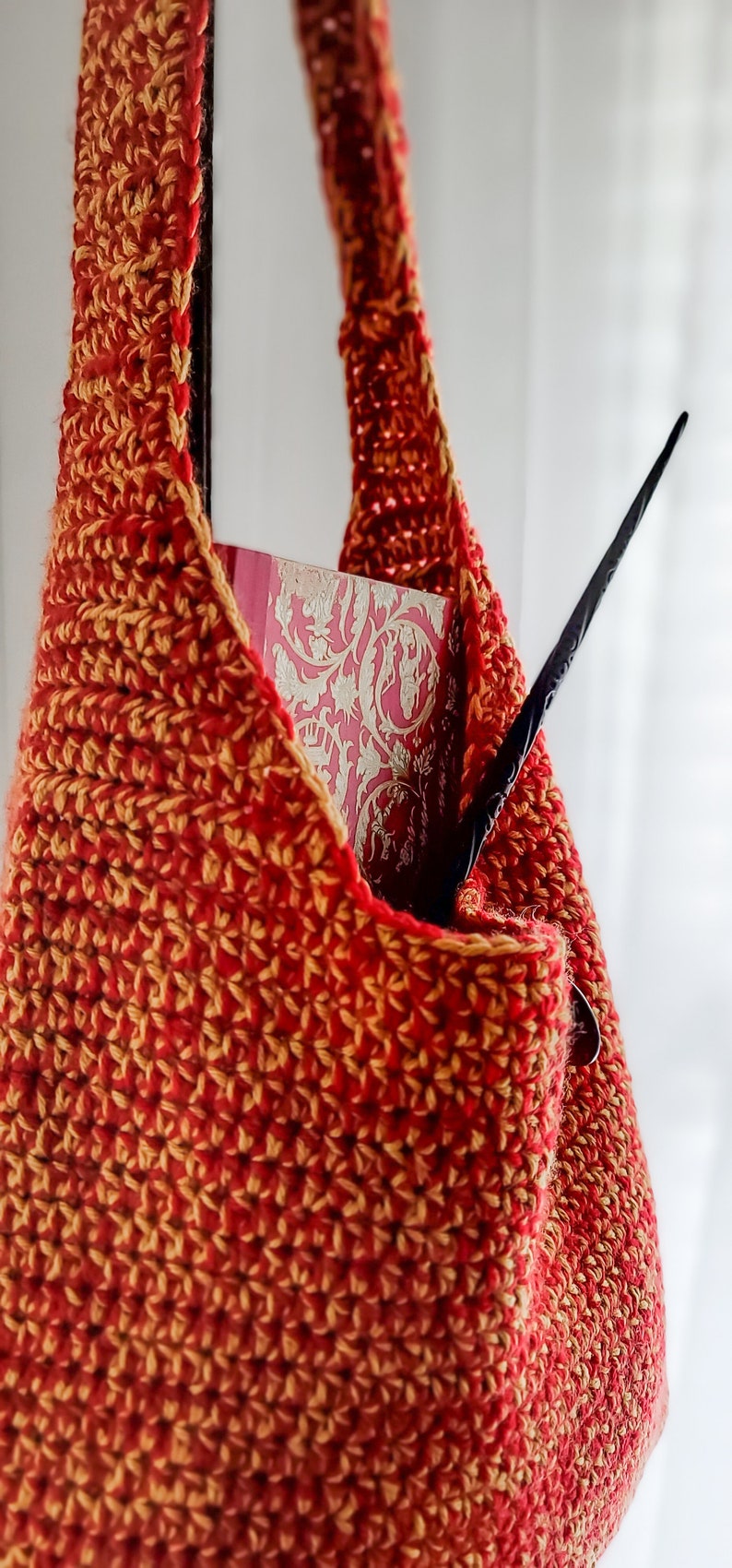 May include: A handmade crochet bag in shades of red and gold, with a long strap. The bag contains a book with a pink cover and a black wand. The bag is made of a textured yarn.
