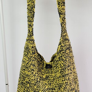May include: A yellow and black crocheted shoulder bag with a single button closure. The bag is suspended from a wooden hanger against a white background. The bag's strap is wide and the body is a large, open tote shape.