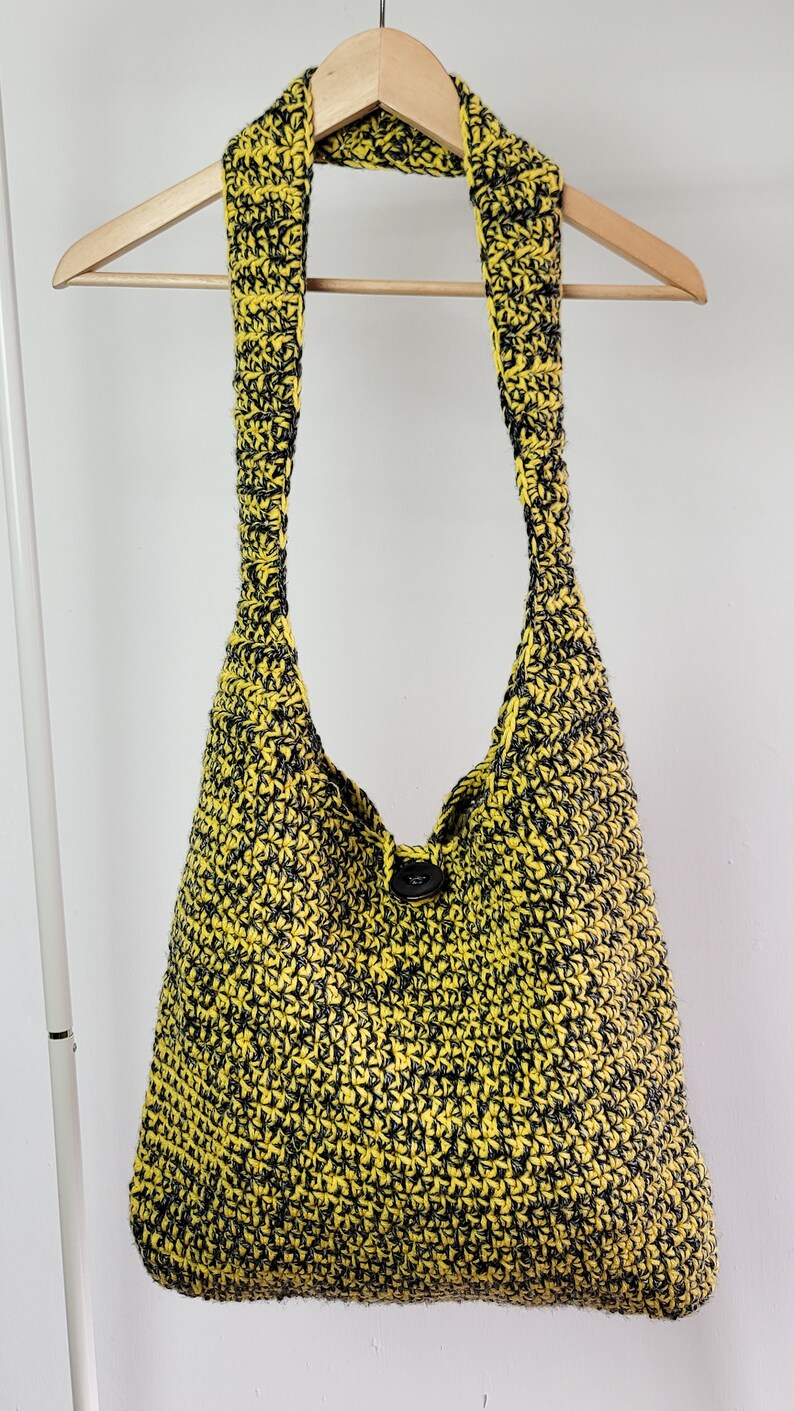May include: A yellow and black crocheted shoulder bag with a single button closure. The bag is suspended from a wooden hanger against a white background. The bag's strap is wide and the body is a large, open tote shape.