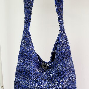May include: A blue and white crocheted shoulder bag with a single black button closure. The bag is made of a textured yarn and hangs from a wooden hanger. The bag's strap is wide and integrated into the body of the bag.