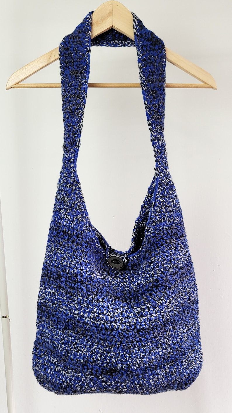 May include: A blue and white crocheted shoulder bag with a single black button closure. The bag is made of a textured yarn and hangs from a wooden hanger. The bag's strap is wide and integrated into the body of the bag.