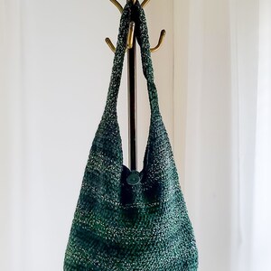 May include: A handmade, green and white crocheted shoulder bag with a button closure. The bag is hanging on a brass coat rack. The bag's texture and color variations create a unique, stylish accessory.