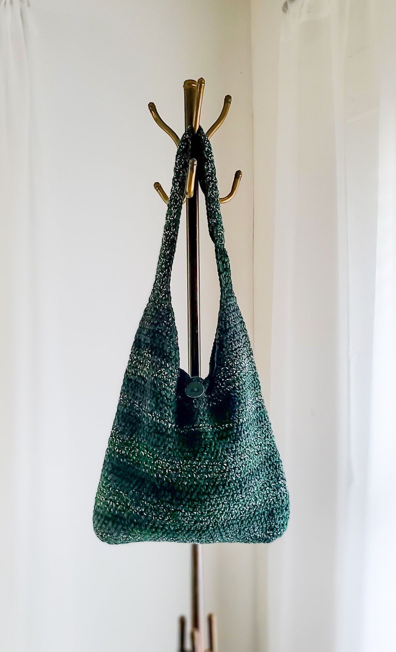 May include: A handmade, green and white crocheted shoulder bag with a button closure. The bag is hanging on a brass coat rack. The bag's texture and color variations create a unique, stylish accessory.