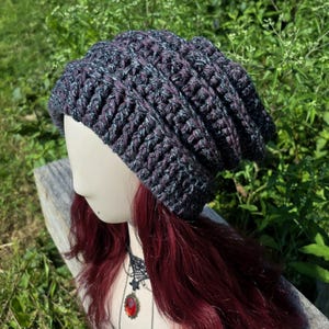 May include: A handmade, slouchy crochet beanie in shades of gray, purple, and blue. The hat is displayed on a mannequin head with long, auburn hair and a black lace choker with a red pendant. The background features green foliage.