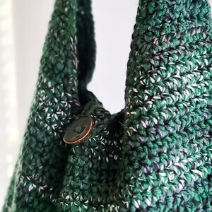 May include: A handmade crochet bag in shades of green, white, and black. The bag features a button closure and a textured stitch pattern. The bag's strap is visible, and the overall design is casual and stylish.