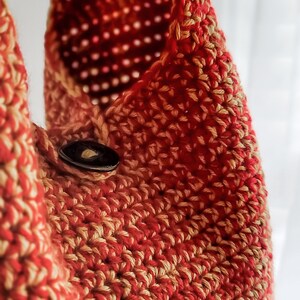 May include: Close-up of a handmade crochet bag in shades of red and tan. The bag features a button closure and a textured, open weave design. The bag is likely made of cotton or a similar yarn.