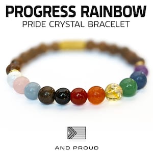 May include: Progress Rainbow Pride Crystal Bracelet with a gold clasp and colorful beads. The bracelet features the colors of the Progress Pride flag. "And Proud" is written below the flag.