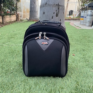 May include: A black rolling suitcase with grey accents rests on green grass. The suitcase has a handle on top and a front pocket with a grey and red design. The brand name "Airzone" is visible on the front.