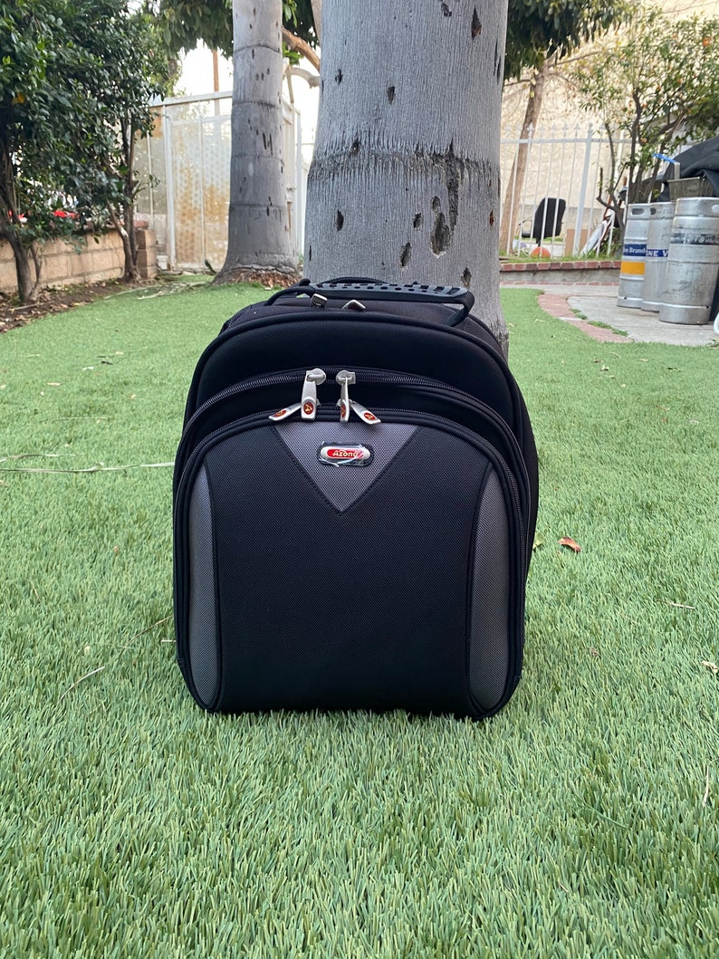 May include: A black rolling suitcase with grey accents rests on green grass. The suitcase has a handle on top and a front pocket with a grey and red design. The brand name "Airzone" is visible on the front.