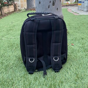 May include: Black backpack with padded shoulder straps, a top handle, and multiple compartments. The backpack is made of a durable material and features silver metal rings for attaching accessories. It is sitting on green grass.